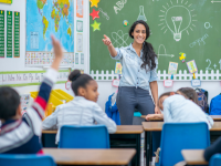 Adult standing at the front of a classroom and chalkboard with kids sitting at school desks